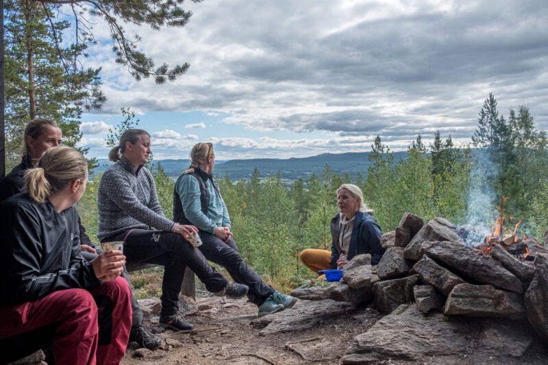 Flera kvinnor sitter framför en eld utomhus under Wise Woman Retreat i Järvsö. Utsikt över Bodasjön. 